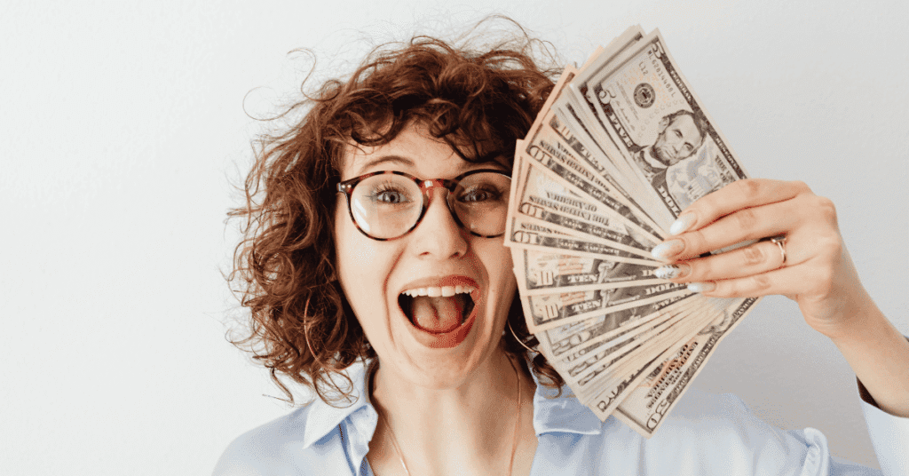 Uma mulher de cabelos cacheados e óculos, com expressão de euforia e boca aberta, segura um leque de notas de dólar em sua mão esquerda. A imagem transmite alegria e sucesso financeiro.
