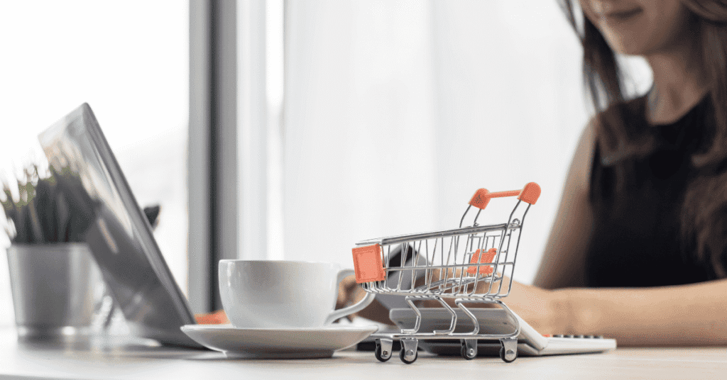 Uma pessoa sentada a uma mesa está usando um laptop, com uma xícara de café e um carrinho de compras em miniatura por perto, sugerindo compras online ou e-commerce. A imagem foca no carrinho de compras e na xícara, com o laptop e a pessoa desfocados ao fundo. Palavras-chave: compras online, e-commerce, carrinho de compras, loja virtual, internet, tecnologia, laptop, café, trabalho remoto, home office.