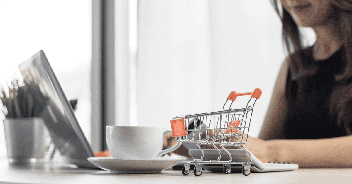 Uma pessoa sentada a uma mesa está usando um laptop, com uma xícara de café e um carrinho de compras em miniatura por perto, sugerindo compras online ou e-commerce. A imagem foca no carrinho de compras e na xícara, com o laptop e a pessoa desfocados ao fundo. Palavras-chave: compras online, e-commerce, carrinho de compras, loja virtual, internet, tecnologia, laptop, café, trabalho remoto, home office.