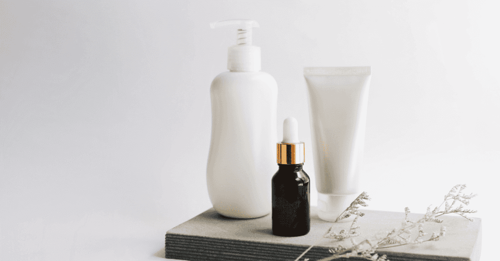 Uma foto minimalista e elegante mostra três produtos de beleza em embalagens brancas e uma garrafa de soro escura com conta-gotas dourado, dispostos sobre uma base de pedra cinza. Ao lado, um delicado ramo seco adiciona um toque natural. A imagem sugere uma rotina de cuidados com a pele ou cabelo, com foco em produtos essenciais.