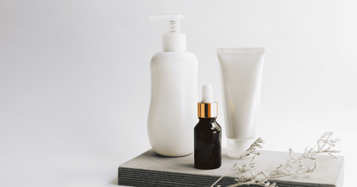 Uma foto minimalista e elegante mostra três produtos de beleza em embalagens brancas e uma garrafa de soro escura com conta-gotas dourado, dispostos sobre uma base de pedra cinza. Ao lado, um delicado ramo seco adiciona um toque natural. A imagem sugere uma rotina de cuidados com a pele ou cabelo, com foco em produtos essenciais.