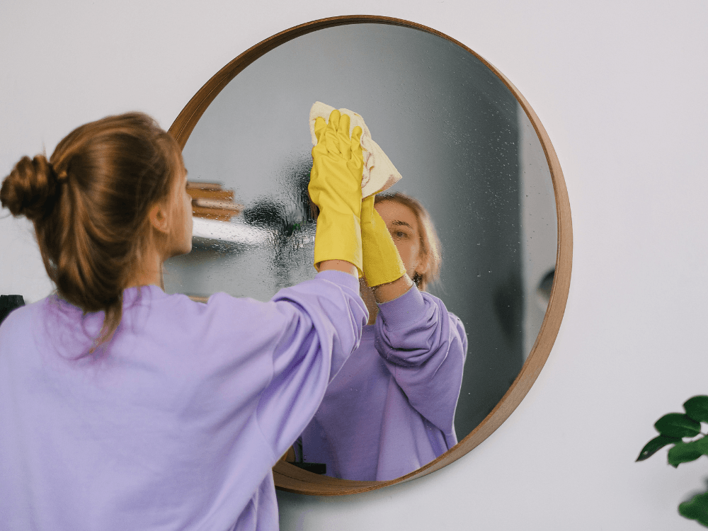 Mulher com luvas amarelas limpando um espelho redondo com um pano, mostrando o uso do vinagre para limpar vidros sem manchas.
