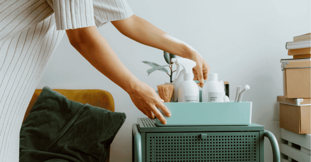 Uma pessoa com um vestido listrado claro segura uma caixa de organização turquesa contendo produtos de limpeza brancos minimalistas, como um spray "MULTI-SURFACE" e um líquido de lavanderia "LAUNDRY LIQUID", junto com uma escova de madeira e outros acessórios. A caixa está sobre um carrinho de metal na mesma cor turquesa, sugerindo uma organização eficiente para limpeza doméstica.