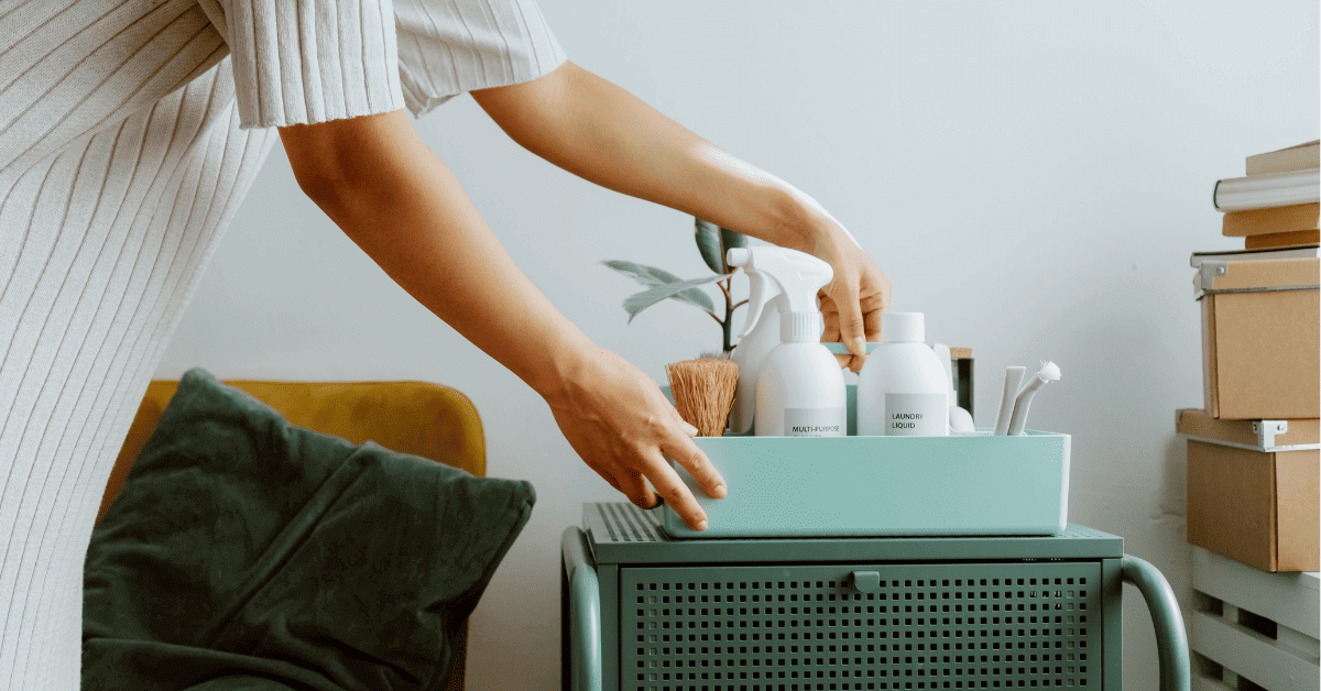 Uma pessoa com um vestido listrado claro segura uma caixa de organização turquesa contendo produtos de limpeza brancos minimalistas, como um spray "MULTI-SURFACE" e um líquido de lavanderia "LAUNDRY LIQUID", junto com uma escova de madeira e outros acessórios. A caixa está sobre um carrinho de metal na mesma cor turquesa, sugerindo uma organização eficiente para limpeza doméstica.