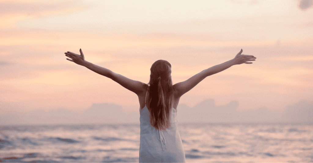 Uma mulher de costas para a câmera, com os braços abertos e levantados em direção a um pôr do sol de tons suaves de rosa e laranja sobre o oceano, as ondas do mar visíveis em primeiro plano.