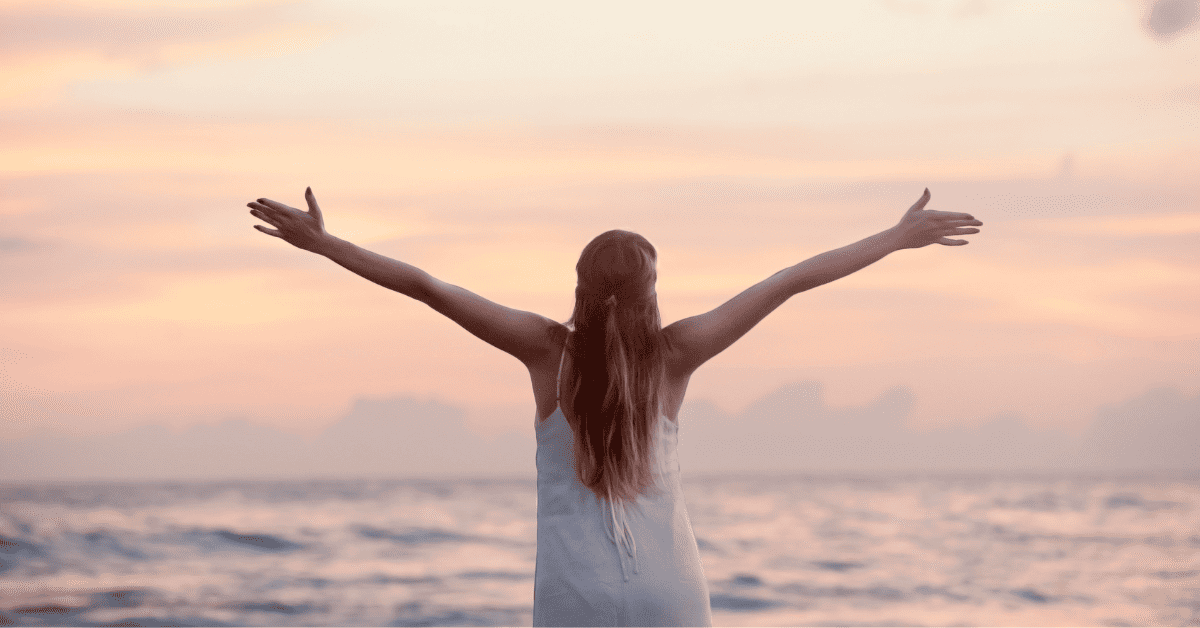 Uma mulher de costas para a câmera, com os braços abertos e levantados em direção a um pôr do sol de tons suaves de rosa e laranja sobre o oceano, as ondas do mar visíveis em primeiro plano.