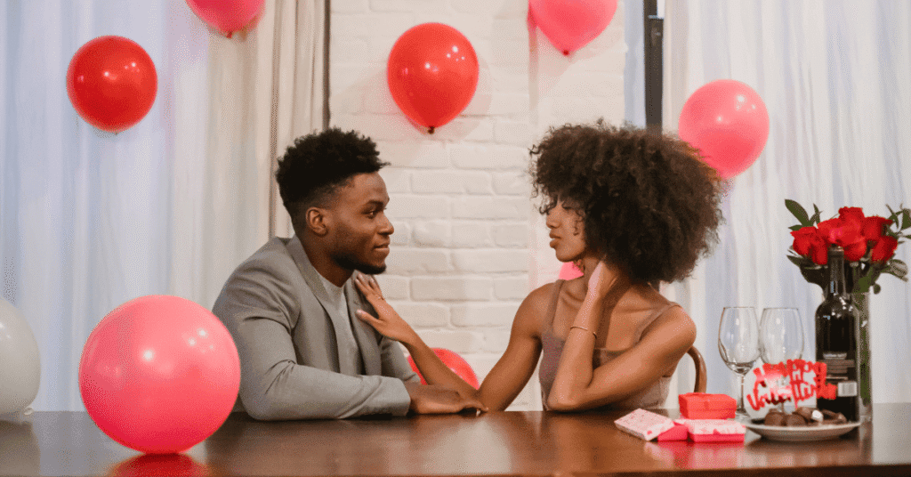 Um jovem casal está sentado frente a frente em uma mesa, olhando um para o outro em um ambiente decorado com balões vermelhos. Há um buquê de rosas vermelhas e dois copos de vinho na mesa.