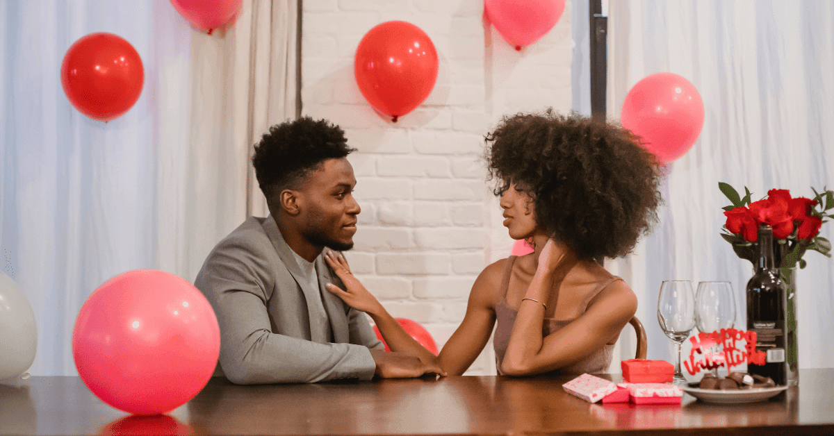 Um jovem casal está sentado frente a frente em uma mesa, olhando um para o outro em um ambiente decorado com balões vermelhos. Há um buquê de rosas vermelhas e dois copos de vinho na mesa.