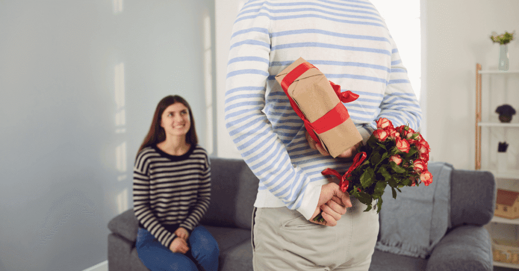 Um homem de costas esconde um buquê de rosas vermelhas e um presente embrulhado atrás das costas, enquanto uma mulher sorridente sentada em um sofá em primeiro plano olha para ele com expectativa