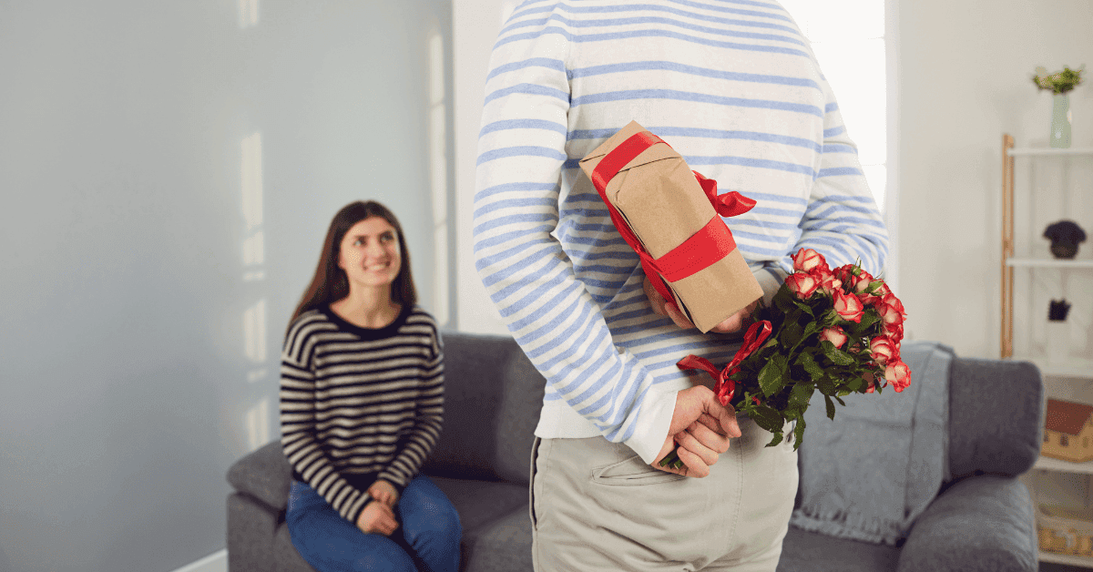 Um homem de costas esconde um buquê de rosas vermelhas e um presente embrulhado atrás das costas, enquanto uma mulher sorridente sentada em um sofá em primeiro plano olha para ele com expectativa