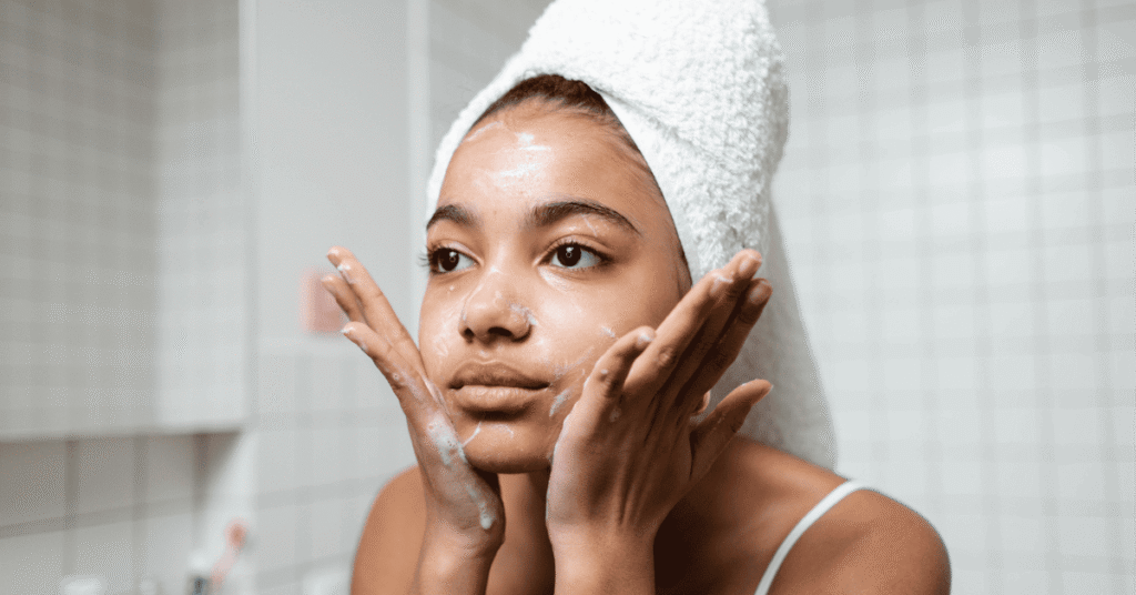 Uma jovem com uma toalha branca enrolada na cabeça aplica um produto de limpeza facial no rosto, com espuma visível na pele