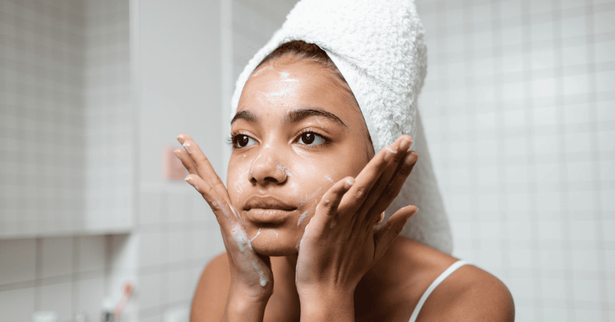 Uma jovem com uma toalha branca enrolada na cabeça aplica um produto de limpeza facial no rosto, com espuma visível na pele